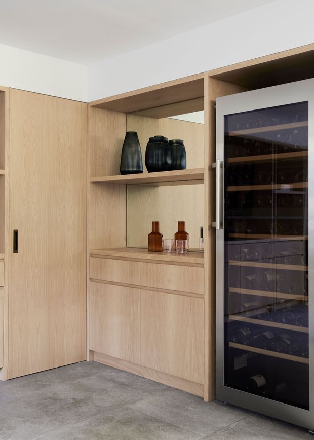 Built-in light timber cabinetry with open display shelving, drawers, and a tall glass-front wine refrigerator. Neutral plaster walls and a grey polished concrete floor create a refined, minimalist storage and wine-display area.