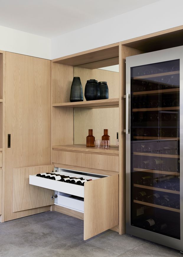 A refined built-in wine storage area with light timber cabinetry, open display shelving, a large glass-fronted wine refrigerator, and a deep pull-out drawer storing bottles. The joinery includes black decorative vases, amber glass bottles, and glasses, set against pale walls and a grey tiled floor.