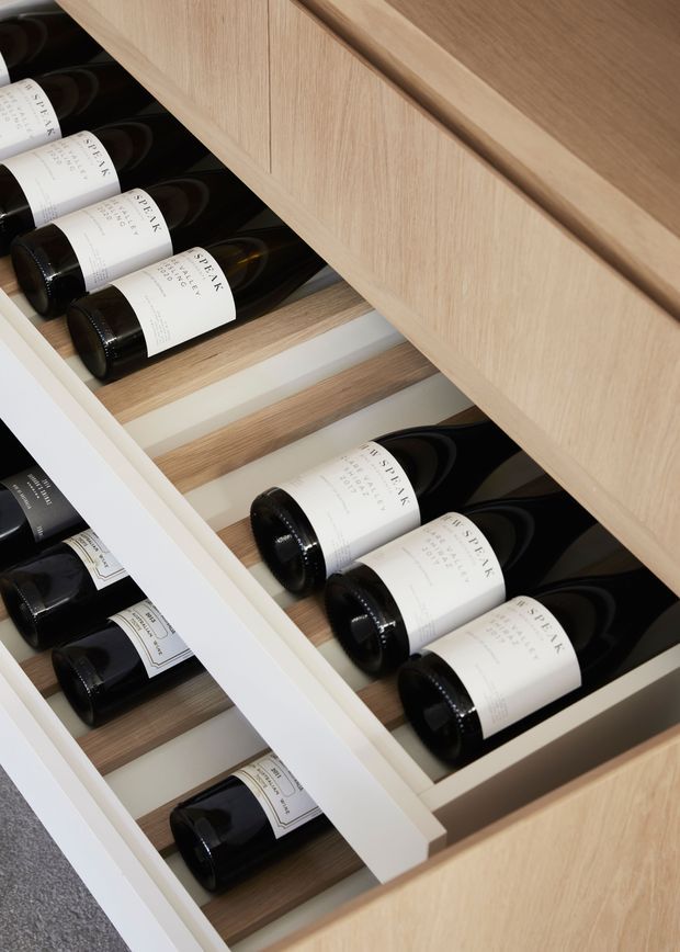 Close-up view of a built-in wine storage drawer with multiple bottles arranged horizontally in individual timber-lined compartments. The joinery has a light natural wood finish with white drawer dividers and surrounding cabinetry, creating a clean, refined storage solution.