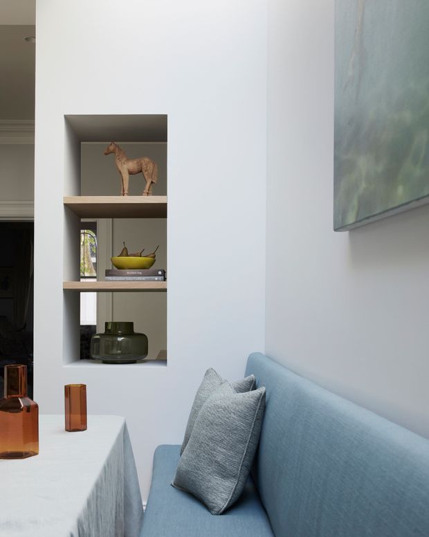 A refined living room corner with a pale blue upholstered sofa, textured blue-grey cushions, and a light table with amber glass vessels. A recessed built-in shelving niche displays books, a ceramic horse sculpture, fruit bowl, and dark green glassware. The walls and joinery are painted white, with a large muted artwork adding subtle colour.