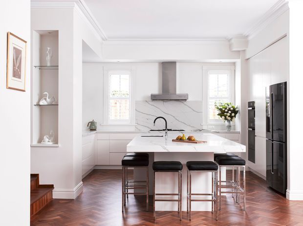 Bright, refined kitchen with a large white stone island and four black leather bar stools, white shaker-style cabinetry, marble-look splashback, integrated appliances, timber herringbone flooring, and a coffered ceiling with detailed cornices. Built-in display niches and classic trim add a traditional character.