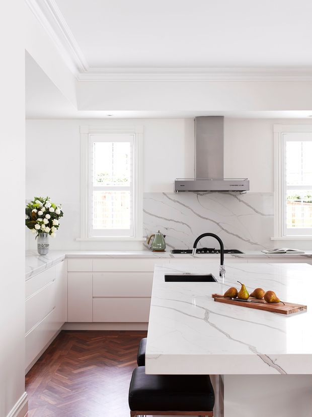Bright, refined white kitchen with a large marble-look island, integrated black sink and curved black tap, white cabinetry, matching splashback, stainless-steel rangehood, and classic white-trimmed windows. A dark herringbone timber floor and black bar stools provide contrast, while fresh flowers and pears add warmth.