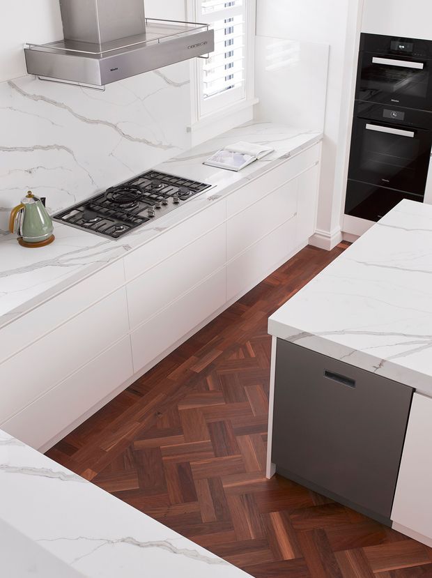 Bright, refined contemporary kitchen featuring white veined stone benchtops and splashback, white panelled cabinetry, a stainless-steel rangehood, black built-in ovens and gas cooktop, and a dark timber herringbone parquet floor. A central island with a dark integrated dishwasher occupies the foreground, while a shuttered window provides natural light.