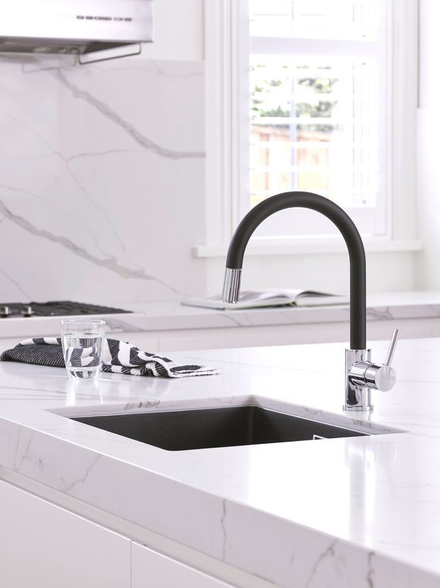 Bright contemporary kitchen detail featuring a white marble-look stone splashback and island benchtop, an undermount black sink, and a tall matte-black and chrome gooseneck mixer tap. White cabinetry, stainless steel rangehood, gas cooktop, and a sunlit window complete the clean, high-contrast composition.