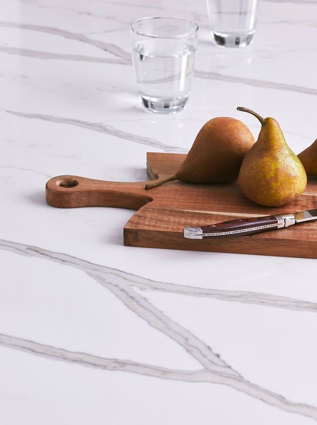 Close-up product-style image of a white marble-look stone benchtop with prominent grey veining. Two wooden cutting boards, pears, clear drinking glasses and a knife are arranged on the surface, demonstrating the countertop finish.