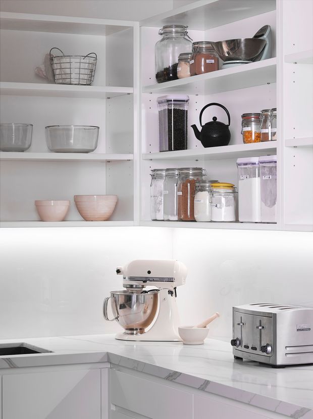 Bright, clean white kitchen with open pantry-style shelving displaying glass storage jars, bowls, cookware and a black kettle. A white stand mixer and stainless-steel toaster sit on a white marble-look benchtop, with white cabinetry and a glossy white splashback. The compact corner layout is softly lit and highly organized.