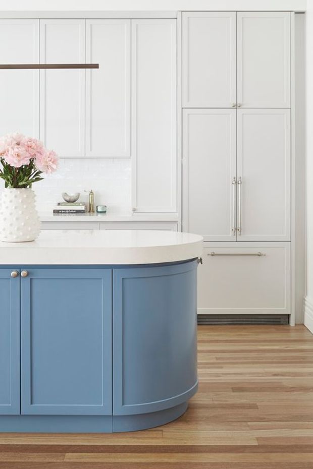Bright, refined kitchen featuring a large curved island with blue painted panelled cabinetry and a white benchtop. The surrounding walls are lined with white and pale grey shaker-style cabinetry, including tall integrated storage, while warm timber flooring adds contrast. Brass-toned hardware, a white tiled splashback, and a vase of flowers complete the elegant traditional scheme.