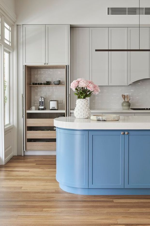 Bright, refined kitchen featuring a large curved blue island with a white benchtop, pale grey-white shaker cabinetry, integrated appliance pantry, glossy light tile splashback, and warm timber flooring. Tall windows provide abundant natural light, while a slim linear pendant adds a contemporary detail.
