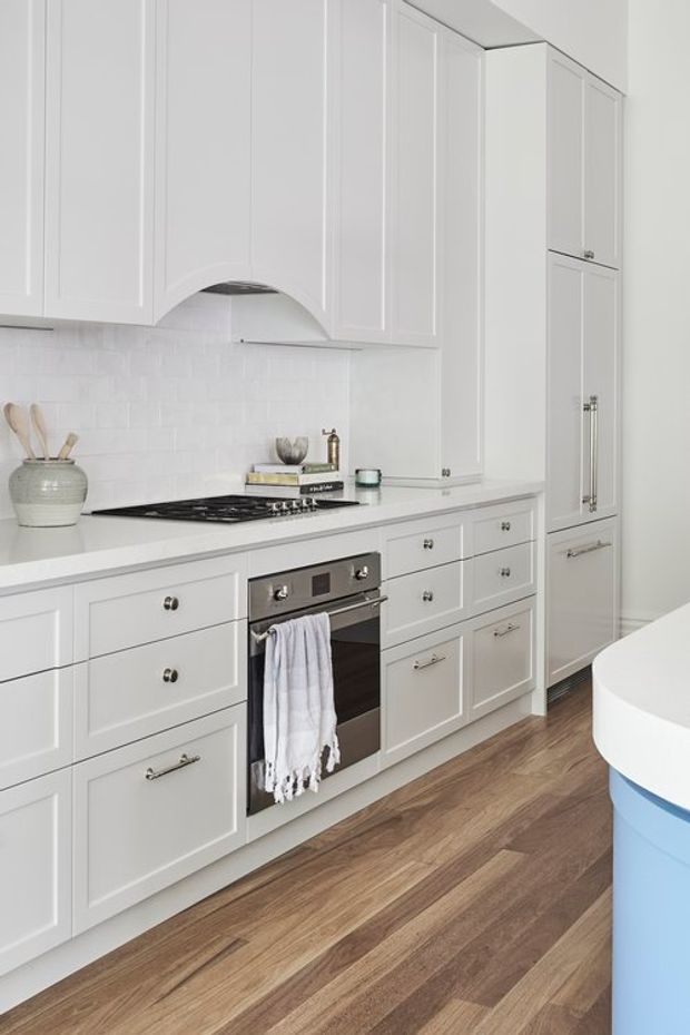 Bright white kitchen with extensive shaker-style cabinetry, a white subway-tile splashback, integrated tall storage, gas cooktop and built-in oven. Warm timber-look flooring contrasts with the white joinery, and a curved island or peninsula edge is visible in the foreground.