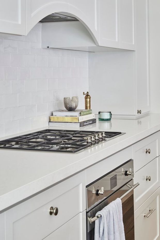 Close-up of a bright, refined white kitchen showing shaker-style cabinetry, a white subway-tile backsplash, pale benchtops, a built-in gas cooktop and stainless-steel oven. An arched decorative rangehood surround, small brass hardware, and restrained countertop accessories add traditional detailing.