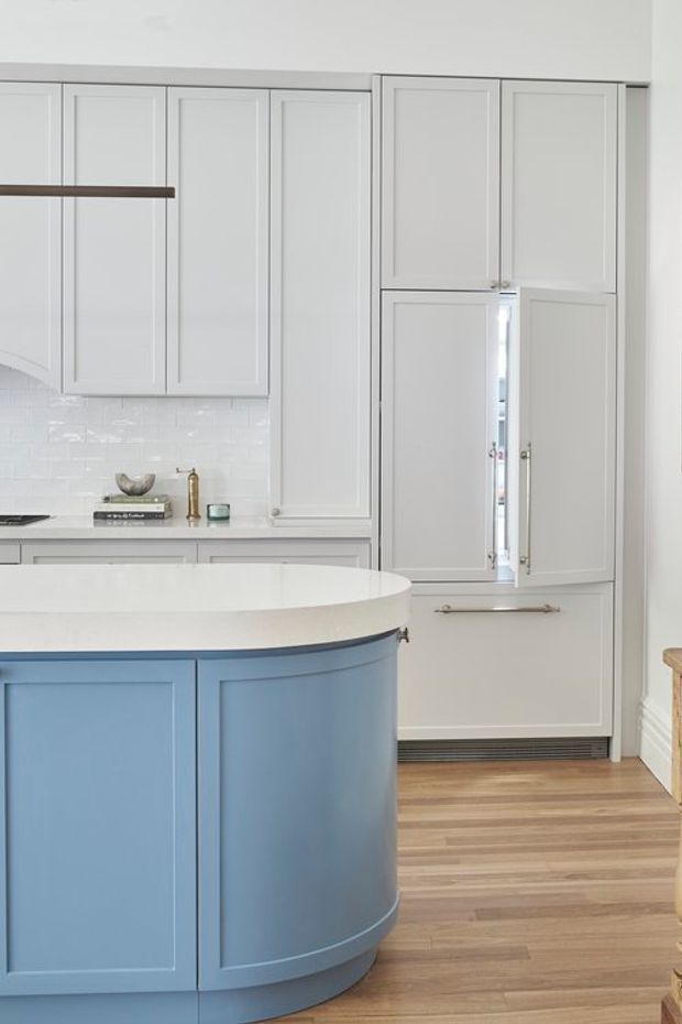 Bright, refined kitchen featuring a large curved blue island with white benchtop, pale grey shaker-style cabinetry, integrated tall storage, white subway-tile splashback, and warm timber flooring. The space has a clean, classic look with subtle brass and dark metal accents.