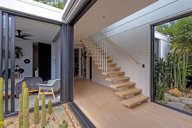 Contemporary open-plan interior centred on a floating timber staircase with slim metal balustrades, set against a white brick feature wall. Large black-framed glazed openings connect the circulation space to a furnished living room on one side and a landscaped courtyard on the other. Light timber flooring, recessed ceiling lights, indoor plants, and a skylight create a bright indoor-outdoor composition.