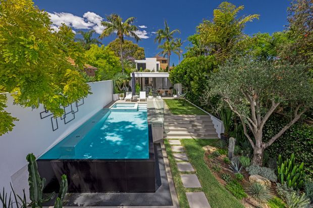 A bright tropical residential backyard viewed from above, featuring a long turquoise swimming pool bordered by white rendered walls and dark pool cladding. A stepping-stone path and broad stairs lead through manicured planting toward a modern multi-level house with outdoor living space. Palm trees, lush hedges, sculptural trees, and strong sunlight create a resort-like setting.