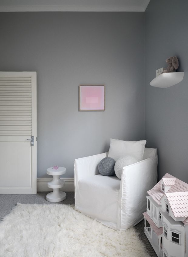 A softly styled children's room corner with a white slipcovered armchair, round cushions, a small white pedestal side table, a plush white rug, and a detailed dollhouse. The room has cool grey walls, white trim, a louvered door, a small pink artwork, and a wall-mounted shelf holding a stuffed toy. The overall look is calm, light, and playful.