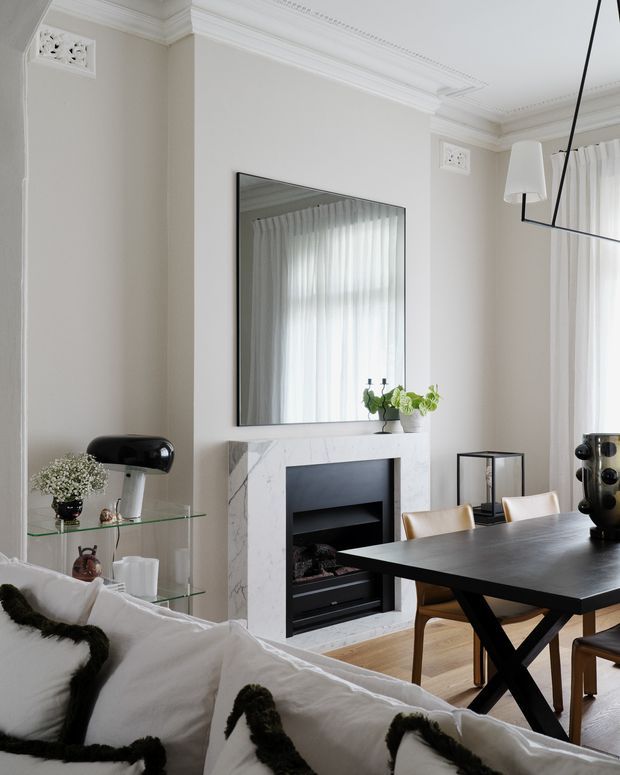 Bright, refined open-plan living and dining interior with classic architectural detailing. The room features ornate white cornices and ceiling mouldings, pale neutral walls, a marble fireplace surround with black inset, a large wall mirror, sheer curtains, a black dining table with timber chairs, and a white sofa in the foreground. A sculptural linear pendant light and small decorative objects add contemporary contrast.