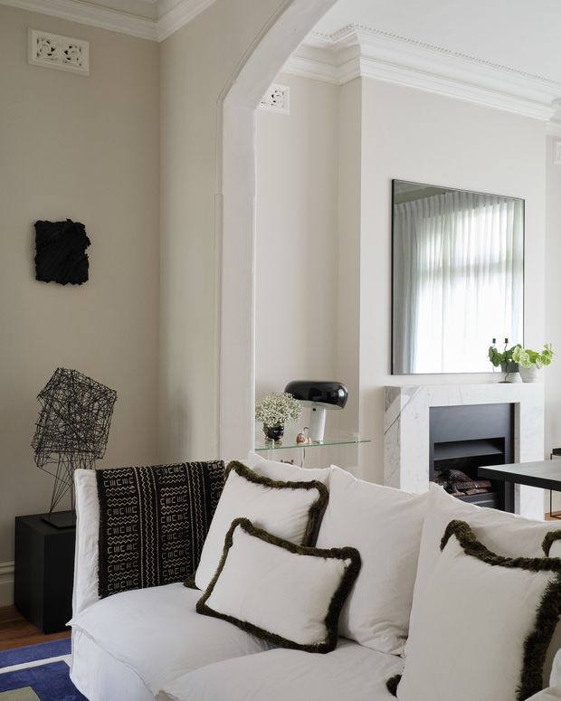 Refined living room interior with a large white sofa layered with white cushions and dark textured throws, set against warm neutral walls. The room features ornate classical architectural detailing, including ceiling cornices, decorative vents, and a broad arched opening. A marble fireplace with a large mirror above it forms the main focal point, while sculptural black decor and a glass console add contemporary contrast.