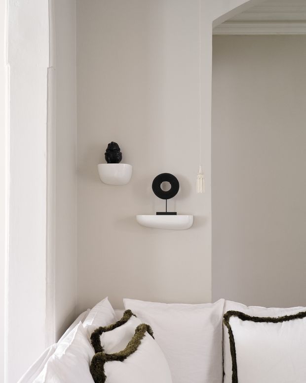A close-up of a softly styled bedroom corner with white bedding and pillows trimmed in dark green textured edging. A pale neutral wall features small floating white shelves displaying minimalist black decorative objects, with traditional layered cornice detailing visible at the upper right and a hanging tassel accent.