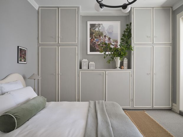 A calm, refined bedroom with a partly visible bed dressed in white bedding and a muted sage bolster cushion. The room features extensive built-in grey cabinetry with white trim and panelled doors, a matching central console, soft grey walls, light carpet, a woven runner, framed artwork, greenery, and a simple ceiling light.