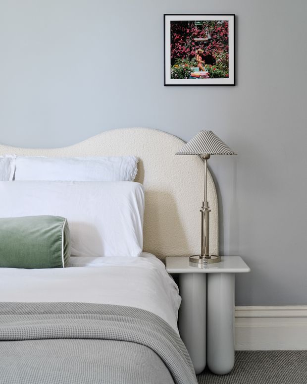 A softly styled bedroom corner with a white upholstered bed and textured curved headboard, crisp white bedding, a muted sage-green accent cushion, and a grey throw. A sculptural light-grey bedside table supports a metallic lamp with a pleated shade. The room has cool grey walls, white skirting boards, grey carpet, and framed botanical artwork.