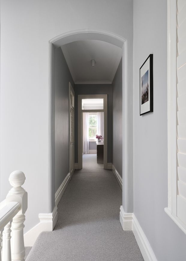 A narrow residential hallway viewed from a stair landing, with grey carpet, pale grey walls, white decorative architraves and skirting, and a curved arched opening leading toward a bright room with a window. A white stair balustrade is visible in the foreground, with framed wall art and louvered doors along the corridor.