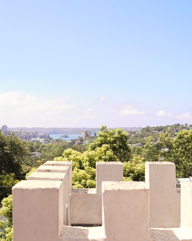 Bright exterior view from an elevated property, looking across leafy residential surroundings toward a distant urban skyline and body of water. The foreground features pale rendered masonry parapet walls with crenellated or stepped detailing, creating a rooftop terrace or balcony-like setting. Clear blue sky and abundant greenery contribute to an airy, sunlit atmosphere.