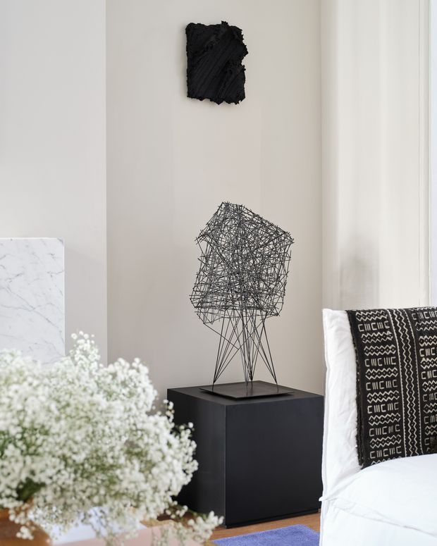 A refined bedroom corner with a neutral beige wall, black sculptural wire artwork displayed on a dark pedestal, and a bed partially visible at right with crisp white bedding and a black-and-white patterned throw. A marble-finish surface and soft white floral arrangement appear in the foreground, creating a calm, contemporary gallery-like setting.