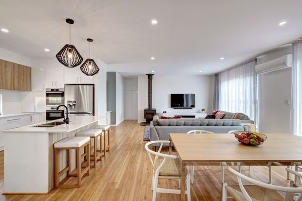 Bright open-plan interior combining a contemporary white kitchen, dining area and living room. The space features light timber flooring, white cabinetry and benchtops, a large island with timber stools, pendant lighting, a timber dining table with white-framed chairs, a grey sectional sofa, a wall-mounted television, and expansive sheer-covered glazing. A wood-burning stove provides a distinctive feature in the living area.