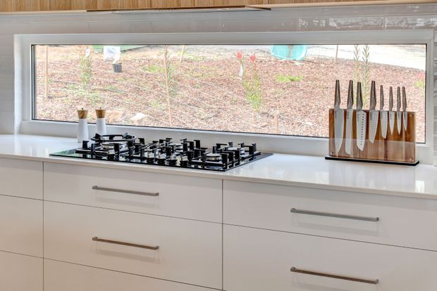 A clean contemporary kitchen work area with a wide horizontal window above a white benchtop. The benchtop contains a black multi-burner gas cooktop, while white handleless-style drawers with slim metal pulls provide cabinetry below. A timber knife block and small countertop accessories sit beside the cooktop. Warm timber wall cabinetry is visible along the top edge, and a landscaped garden is seen outside through the window.