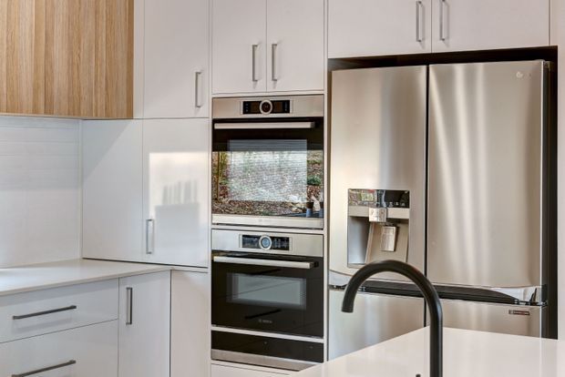Bright contemporary kitchen featuring white full-height cabinetry, timber-grain overhead cabinets, integrated stainless-steel double ovens, a large stainless-steel French-door refrigerator, white tiled splashback, and a curved matte-black tap over a light benchtop.