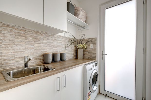 Compact, bright laundry room with white cabinetry, a timber-look benchtop, stainless-steel sink, front-loading washing machine, textured beige tile splashback, open shelving, and a frosted-glass external door. Neutral finishes and simple styling create a clean, practical space.