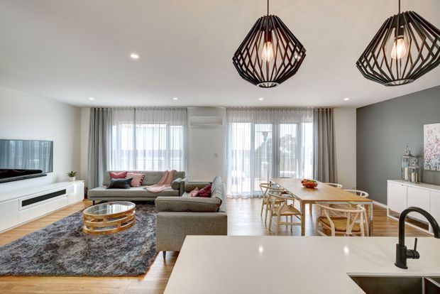 Open-plan contemporary living, dining and kitchen area with light timber flooring, white cabinetry and benchtops, a grey feature wall, large sheer-covered windows, and two prominent geometric black pendant lights. The living zone includes a grey sectional sofa, textured rug and round timber coffee table, while the dining area has a timber table and chairs. A kitchen island with a black gooseneck tap occupies the foreground.
