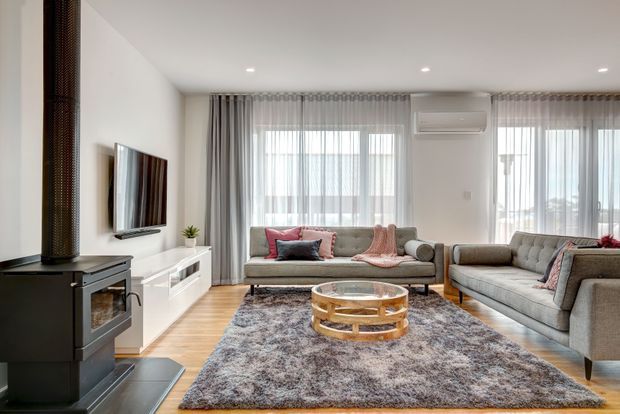 Bright contemporary living room with two grey upholstered sofas arranged around a round glass-and-wood coffee table. The room features light timber flooring, a large textured rug, sheer floor-to-ceiling curtains, expansive windows, white built-in media cabinetry, recessed ceiling lights, and a black freestanding wood-burning fireplace.