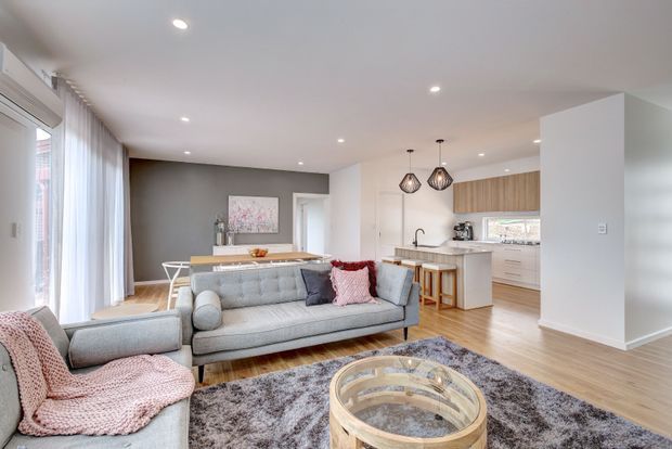 Bright open-plan living, dining and kitchen area with light timber flooring, soft grey furnishings, a textured grey rug, white walls and cabinetry, and warm timber accents. The living room features a large sofa and armchair, while the kitchen includes a peninsula with stools, pendant lights and integrated cabinetry. Sheer curtains and recessed lighting create an airy contemporary interior.