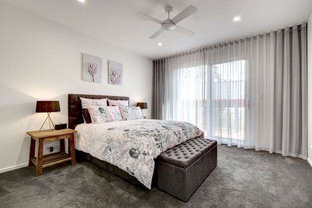 Bright, contemporary bedroom with a large upholstered bed dressed in pale floral bedding, matching botanical wall art, dark timber bedside tables, a tufted upholstered bench at the foot of the bed, grey carpet, and full-height sheer and blackout curtains. A ceiling fan and recessed lighting complete the clean, comfortable interior.