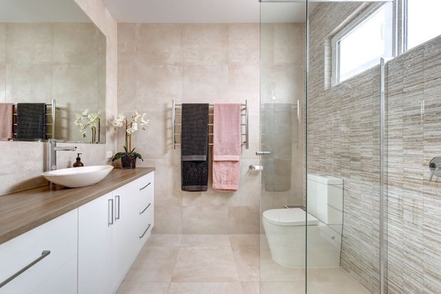 A bright contemporary bathroom with a long white floating vanity, timber-look benchtop, vessel basin and wall-mounted tapware. Large beige stone-look tiles cover the floor and walls, while a textured horizontal feature tile lines the shower wall. The room includes a glass shower enclosure, freestanding toilet, mirrored wall, heated towel rails with dark and blush towels, and a high horizontal window.