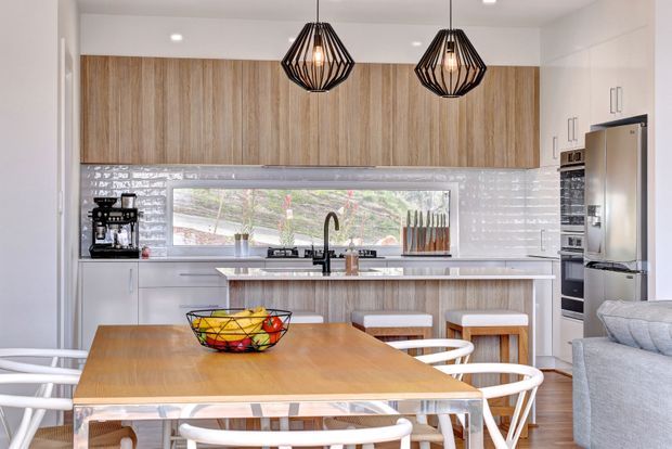 Bright open-plan kitchen and dining area with warm vertical timber cabinetry, glossy white subway-tile splashback, white island bench, stainless-steel appliances, and a timber dining table with white chairs. Black geometric pendant lights provide a strong decorative accent, while a large horizontal window brings in natural light and overlooks greenery.