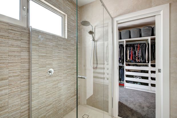 Bright contemporary shower room with a frameless glass enclosure, wall-mounted shower fixtures, large horizontal window, and textured beige stone-look wall tiles. An open doorway reveals a carpeted walk-in wardrobe with extensive white built-in shelving and hanging storage.