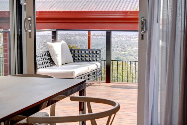 Bright view from an interior dining area through open glazed doors onto a timber deck or balcony. The outdoor space has a woven bench with white cushions, black metal balustrading, and a red overhead awning or roof element, with a distant urban landscape beyond. A dining table and wooden chair are visible in the foreground.