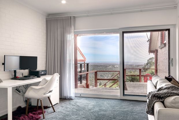 Bright compact living room and home office nook with a wall-mounted television, white desk, swivel chair, sofa, and large sliding glass doors opening onto a timber balcony with expansive coastal views. The room features white painted brick walls, pale grey carpet, sheer curtains, and a neutral contemporary fit-out.