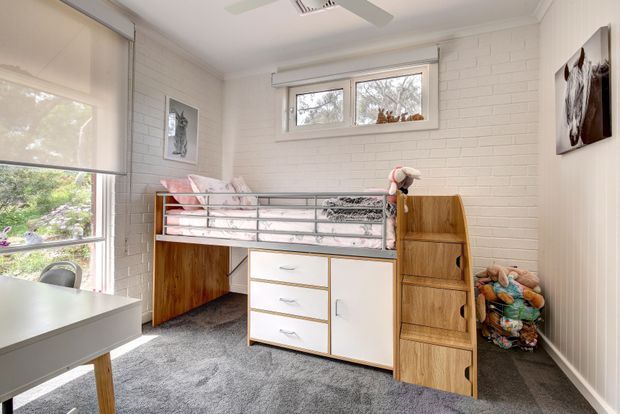 Bright children's bedroom with a built-in loft-style bed and integrated timber-and-white storage drawers, shelving and wardrobe. The room has white-painted brick walls, grey carpet, roller blinds, a ceiling fan and large windows overlooking greenery. Plush toys and soft bedding add a playful, lived-in character.