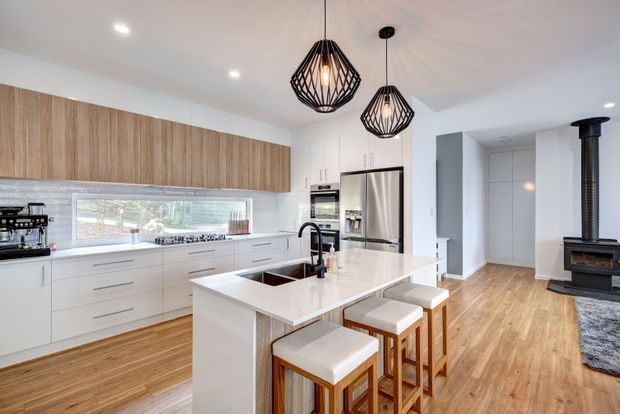 Bright, spacious contemporary open-plan kitchen with white cabinetry, a large white island with seating for four, integrated stainless-steel appliances, and a warm timber floor. A timber-look overhead cabinet bank adds warmth, while black geometric pendant lights and a black wood-burning stove provide contrast. The kitchen opens toward a hallway and adjoining living area.