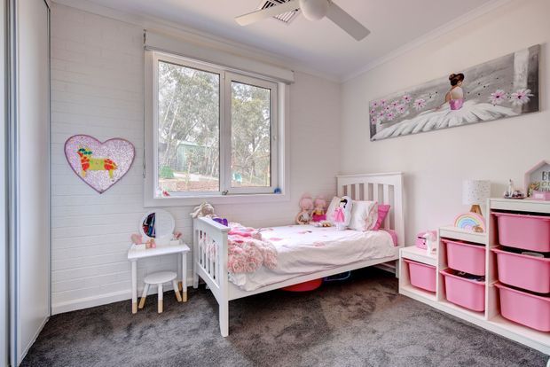 A bright, playful children's bedroom with a white-painted horizontal wall lining, grey carpet, and white furniture. The room contains a single white bed dressed in pink and white bedding, a small children's desk and chair, pink storage bins in open shelving, and colourful child-oriented artwork and toys. A large window provides natural light, and a ceiling fan is fitted overhead.
