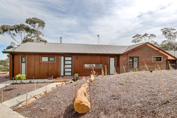 Single-storey residential home viewed from the front, combining vertical timber wall cladding with a dark red-brown brick gabled wing and a low-pitched corrugated metal roof. The facade includes a glazed entry door, horizontal windows, and a landscaped gravel garden with stone edging and native trees in the background.