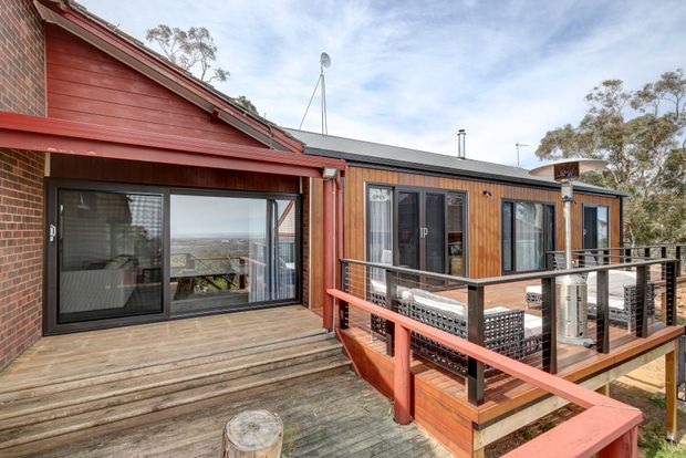 Exterior view of a timber-clad, multi-level residential dwelling with a red-painted deck and balustrade. Large glazed sliding doors open onto the deck, with a forested setting and distant landscape visible beyond. The building combines warm timber boards, brickwork, metal-framed glazing, and a pitched roof section.