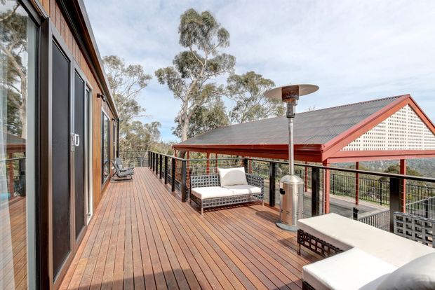 A spacious elevated timber deck beside a contemporary timber-clad building, furnished with outdoor lounge seating and a tall patio heater. Black metal balustrades frame views of eucalyptus trees and a neighbouring pitched-roof structure with red trim and a perforated gable screen. The setting is bright and open, with warm timber surfaces and a relaxed outdoor entertaining feel.