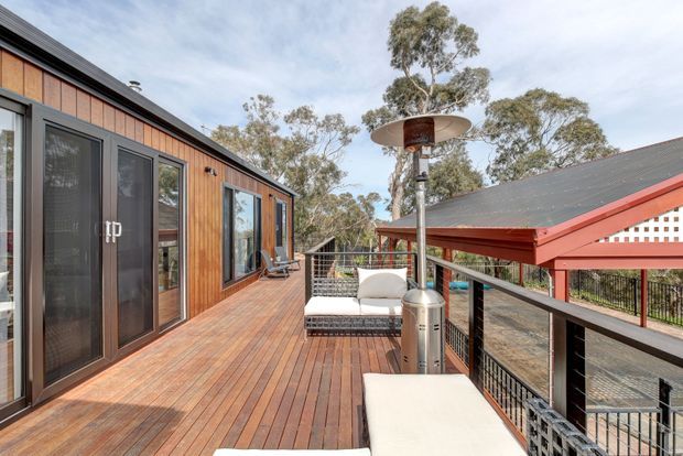 A spacious elevated timber deck and balcony overlooking a wooded setting. The deck is attached to a contemporary timber-clad building with dark-framed glazing, outdoor lounge furniture, a freestanding patio heater, and black metal railings. A red-roofed covered outdoor structure is visible alongside the deck.