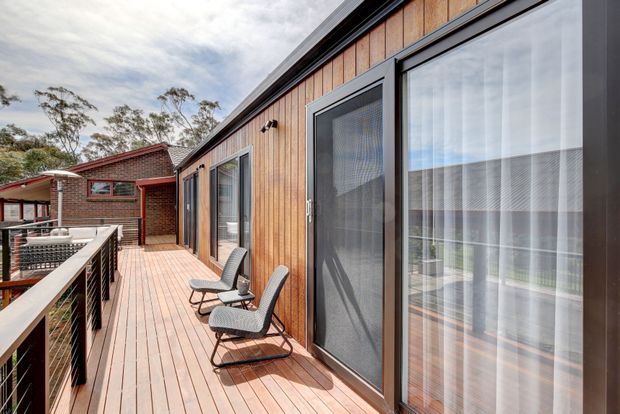 A sunny elevated timber deck running alongside a contemporary building, with large glazed sliding doors and dark-framed windows opening to the interior. The facade is clad in vertical warm timber boards, while a metal balustrade borders the deck. Outdoor lounge chairs are arranged along the walkway, and a brick building with a covered outdoor area is visible beyond.