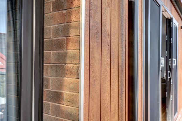Close-up exterior view of a contemporary building facade showing warm red-brown brickwork, vertical timber cladding, and dark aluminium-framed glazing and entry doors. The image focuses on the contrast between masonry, timber, and dark metal elements.