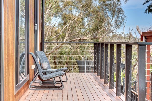 A timber-clad outdoor deck or balcony beside full-height glazed doors, furnished with two reclining chairs and overlooking a wooded landscape. The deck has timber decking boards and a dark metal-and-wire balustrade, with warm natural tones and abundant greenery beyond.