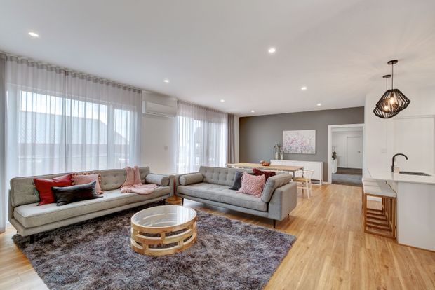 Bright open-plan living and dining area with a contemporary neutral interior. Two light-grey sofas face a round timber coffee table over a large textured rug, while sheer curtains span the windows. The adjoining dining area and white kitchen with island are visible, along with recessed ceiling lights and a black geometric pendant.